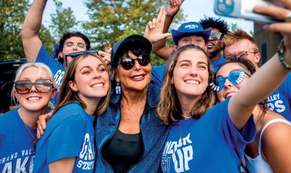 President Mantella poses with a group of students for a selfie at the Grand Valley tailgate. Students wear blue laker gear and Mantella wears a baseball cap and sunglasses.